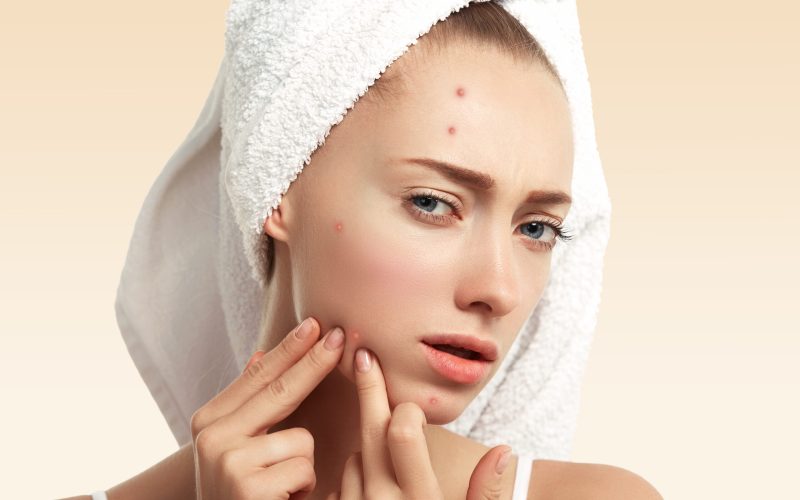 Close up isolated portrait of young woman squeezing pimple on her cheek in the bathroom. Cropped view of Caucasian female having skin problem, against blue wall background. Beauty and spa concept