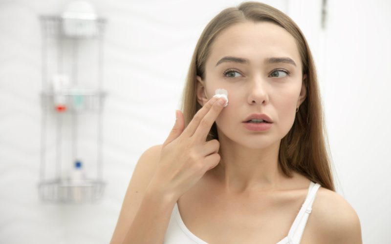 Young beautiful woman applying cream to her face and looking worried at the mirror at home bathroom. Beauty, skin care concept, lifestyle