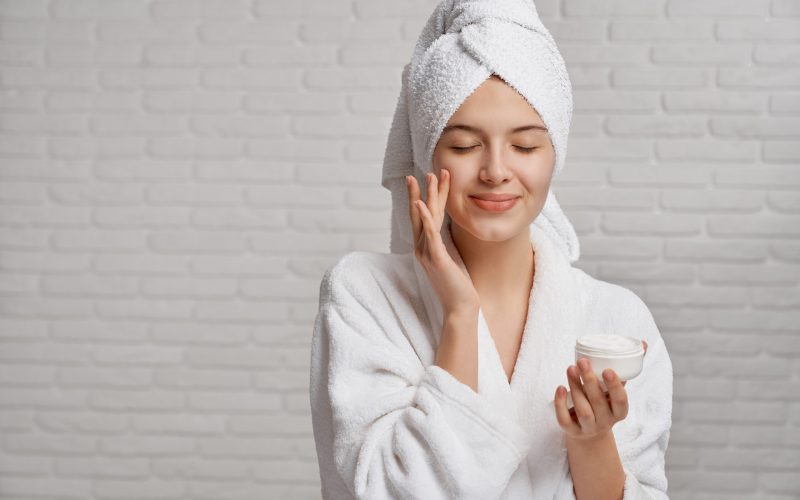 Young, pretty woman with pleasure applying moisturizer on face with closed eyes. Beautiful model in white bathrobe and towel on head posing on grey background. Concept of natural cosmetics.